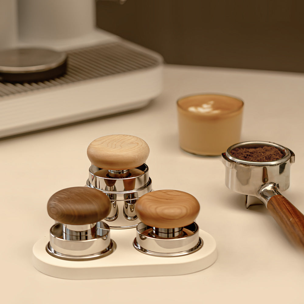 Set of three espresso tampers with wooden knobs on a white surface, with a blurred coffee machine in the background.